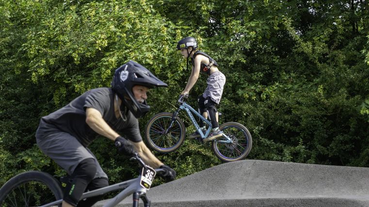 Two cyclists are performing acrobatic tricks on a trail. The surroundings are green and it is a sunny day.