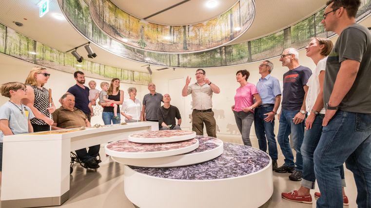 Eine Gruppe von Menschen steht in einem modernen Raum und hört einem Vortrag zu. Im Mittelpunkt befindet sich eine runde Präsentationsfläche.