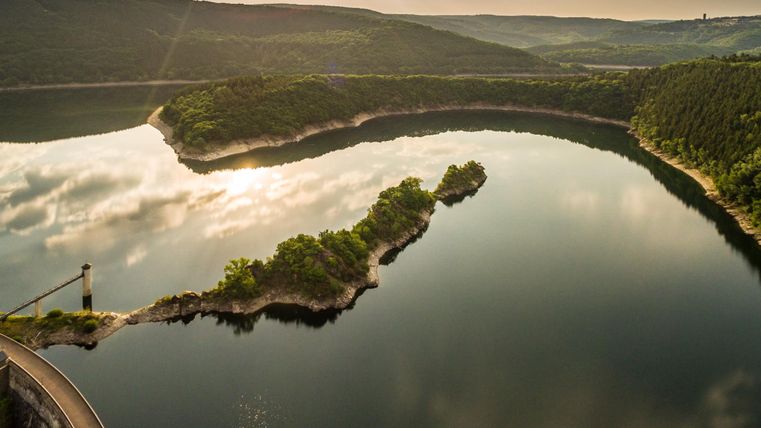 Eine ruhige Seenlandschaft mit sanften Hügeln und einigem Grün. Der Himmel spiegelt sich klar im Wasser.