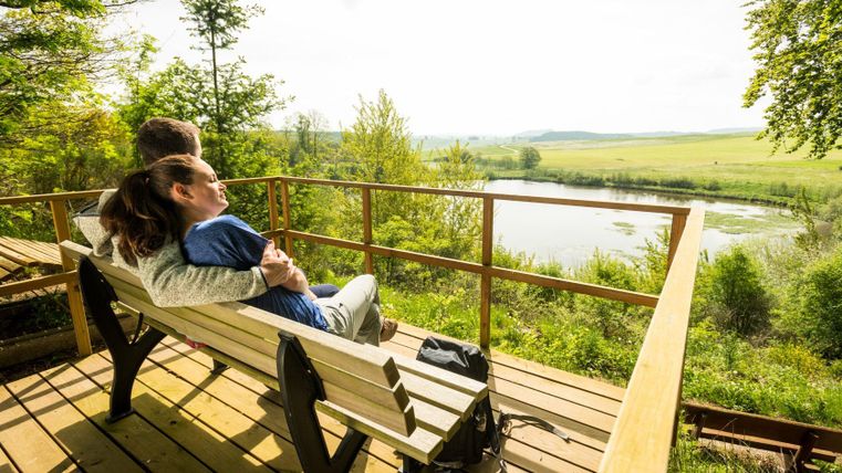 Ein Paar sitzt auf einer Bank und genießt den Blick auf das Eichholzmaar in einer grünen Landschaft.