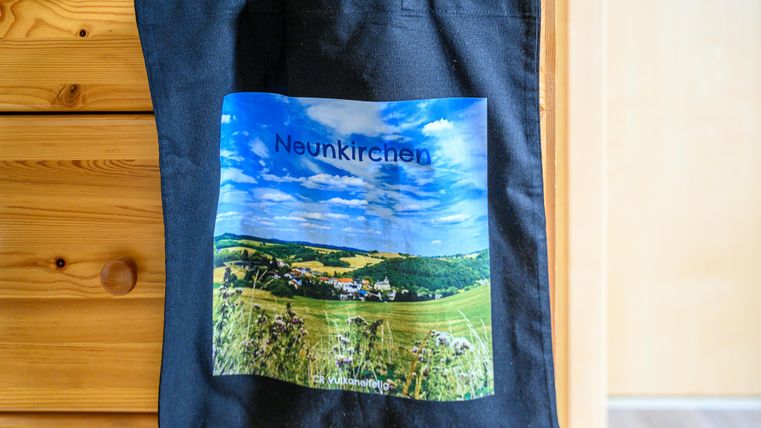 Eine schwarze Tasche mit einem Bild von Neunkirchen. Die Landschaft zeigt Hügel, Wiesen und einen blauen Himmel.