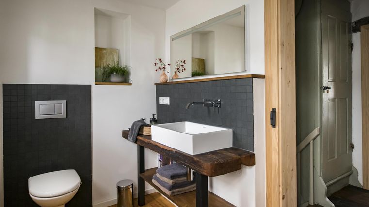 Ein modernes Badezimmer mit einem Waschbecken auf einem Holztisch und einem eleganten Spiegel. Die Wände sind teilweise in dunklem Grau gehalten, und es gibt eine Tür zu einem weiteren Raum.