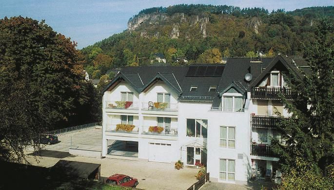A modern building with several floors and balconies. In the background, a wooded slope can be seen.