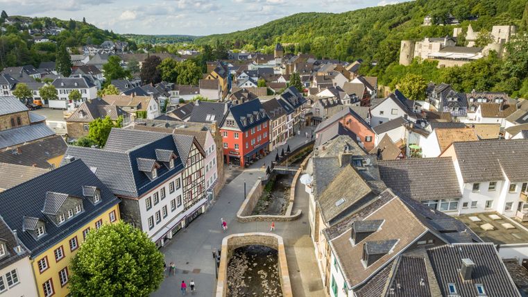 Eine malerische Stadtansicht mit bunten Häusern und einem kleinen Fluss. Im Hintergrund sind grüne Hügel und eine Burg zu sehen.
