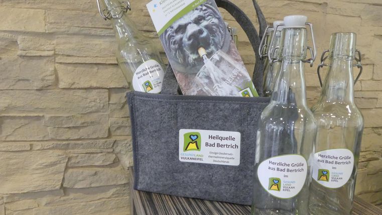 A bag with several glass carafes and a brochure about the healing spring Bad Bertrich. The bottles are labeled and sit on a wooden surface.