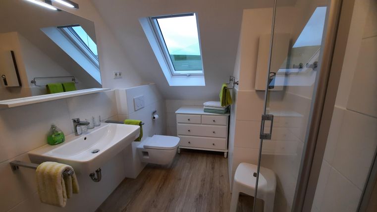 A modern bathroom with a shower cabin and a large window. It is bright and minimally furnished with wooden flooring and green accents.