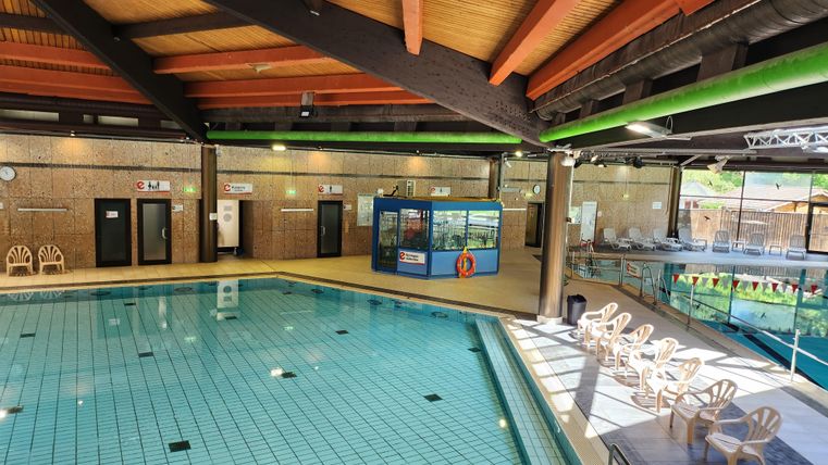 Een binnenzwembad met kristalhelder water en houten plafonds. Aan de zijkant staan comfortabele stoelen en een klein aquariumgebied is zichtbaar.