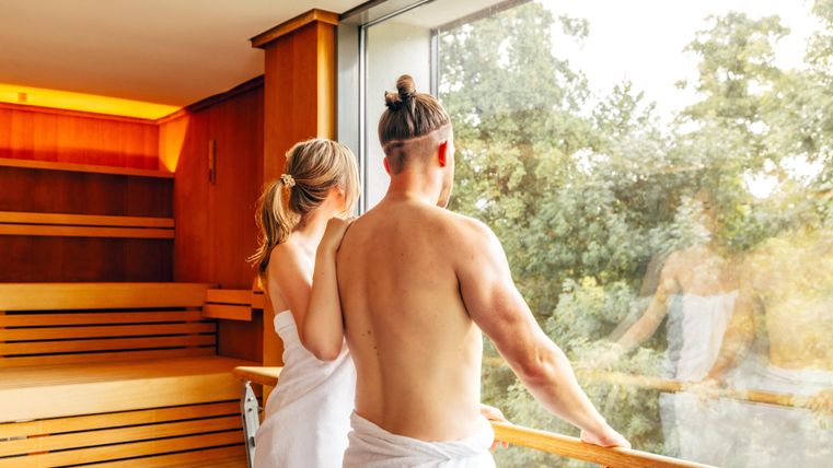 Een paar in saunadoeken staat bij het raam en kijkt naar buiten. De ontspannen sfeer wordt benadrukt door het houten interieur en de groene natuur op de achtergrond.