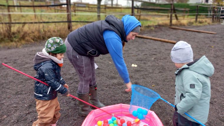 A woman is playing with two children outside. They are catching colorful balls from a pink pool.