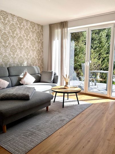 A cozy living room with a gray sofa and a small table. Large windows allow plenty of light in and provide a view of the greenery.