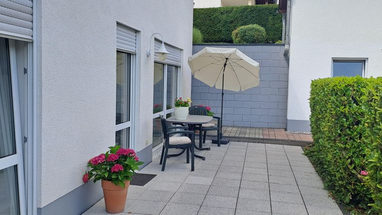 Een gezellige terrasse met een tafel, stoelen en een parasol. Kleurrijke bloemen in een pot complementeren de buitenruimte.