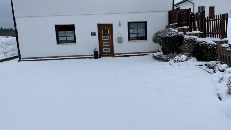 Een huis in de sneeuw met een helder, wit erf. Enkele planten zijn zichtbaar en er staat een hek op de achtergrond.