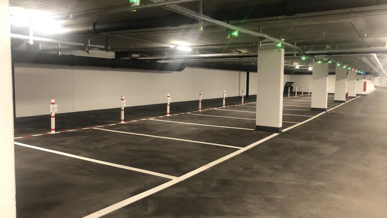 An empty parking garage with several parking spaces. The lighting is bright and there are white markings and traffic bollards.