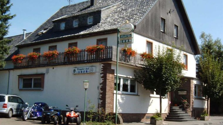 <p>Ein dreigeschossiges Gebäude mit schwarzem Dach.Den Eingang erreicht man über eine Treppe. Im ersten Stock umrahmt ein Holzgeländer mit Blumenkästen einen großen Balkon. Links neben dem Haus ist ein Parkplatz, der mit drei Autos belegt ist. Hinter dem Parkplatz hängt ein Schild, mit der Aufschrift "Biergarten". Vor dem Hotel verläuft ein Bürgersteig, der direkt an die Hauptstrasse angrenzt.</p>