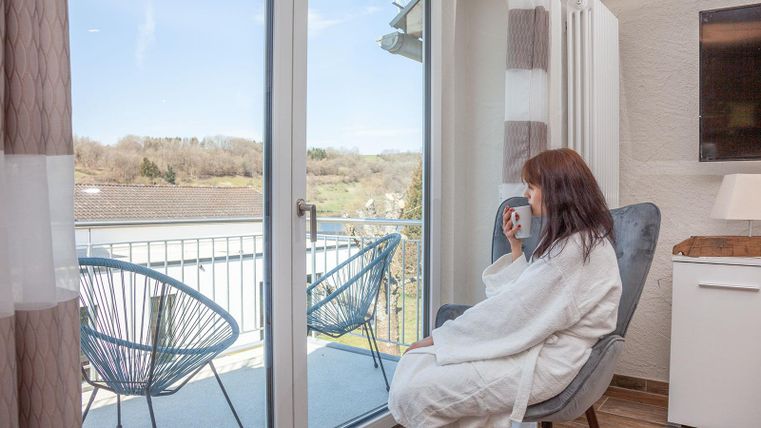 Eine Frau in einem Bademantel sitzt auf einem Stuhl und genießt die Aussicht auf einen Balkon. Im Hintergrund sind zwei Stühle und eine schöne Landschaft zu sehen.