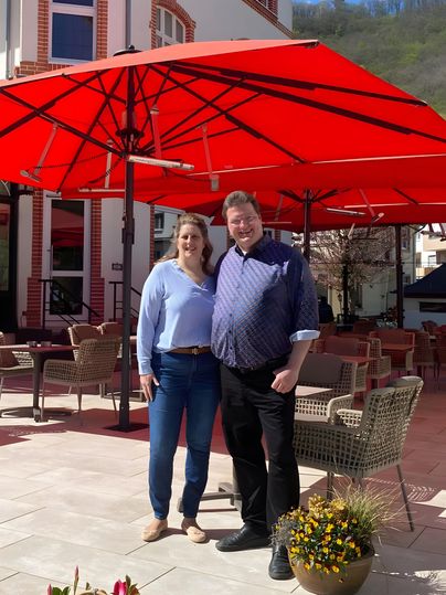 Ein Paar steht lächelnd unter einem großen roten Sonnenschirm. Im Hintergrund sieht man eine gemütliche Terrasse mit Sitzgelegenheiten.