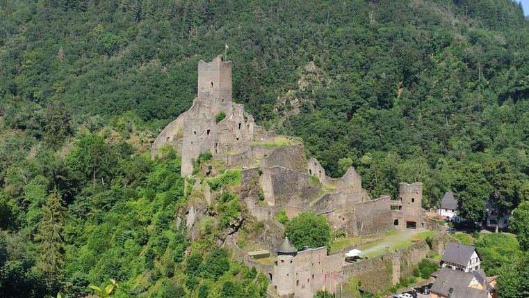 Eine beeindruckende Burgruine auf einem grünen Hügel umgeben von Wäldern. Im Vordergrund sieht man ein kleines Dorf mit typischen Häusern.
