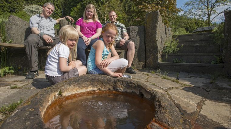 Eine Familie sitzt um eine runde Quelle in einem Garten. Die Kinder beobachten das Wasser, während die Erwachsenen entspannt im Hintergrund sitzen.