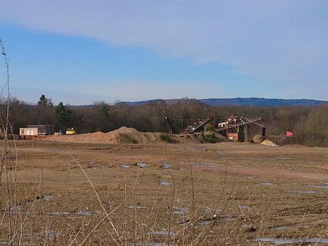 Eine Baustelle mit Erde und Baumaschinen in der Landschaft. Im Hintergrund sind Bäume und ein blauer Himmel zu sehen.