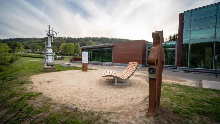 Ein moderner Außenbereich mit einer Skulptur, einer Bank und einem großen Fensterbau. Im Hintergrund ist eine grüne Landschaft sichtbar.