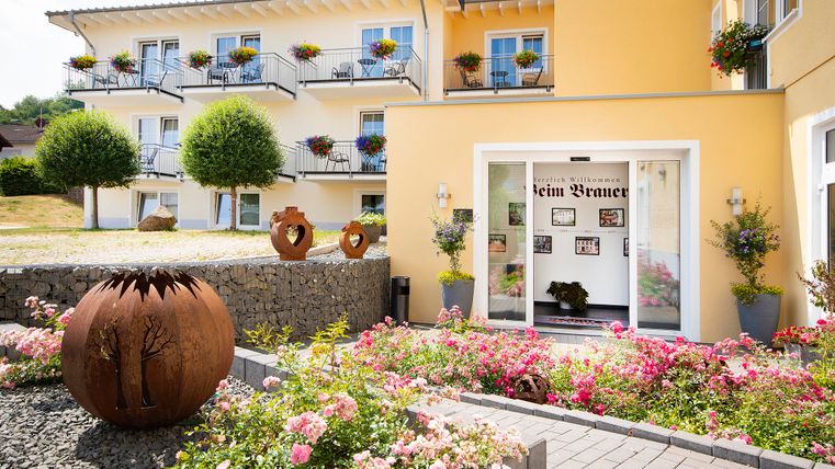 An inviting entrance area with colorful flowers and a well-maintained garden. In the background, a bright, multi-story building can be seen.