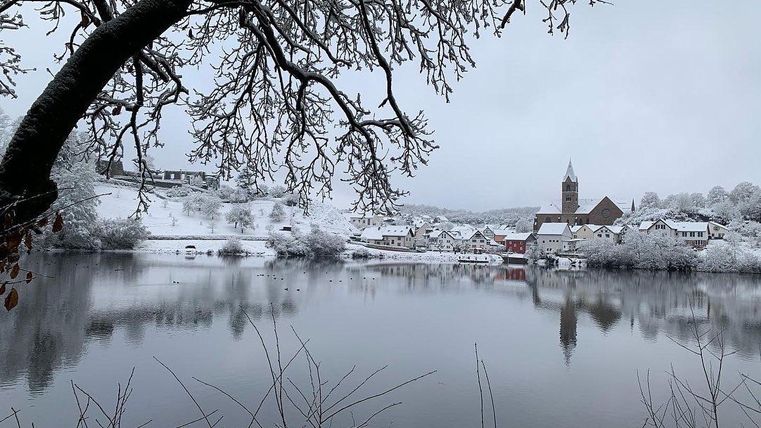 A wintry landscape with snow-covered trees and a calm lake. In the background, some houses and a church tower are visible.