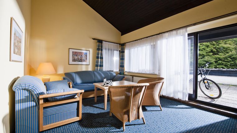 Ein komfortabler Wohnbereich mit blauen Möbeln und einem Tisch. Große Fenster lassen viel Tageslicht herein und bieten einen Blick nach draußen.