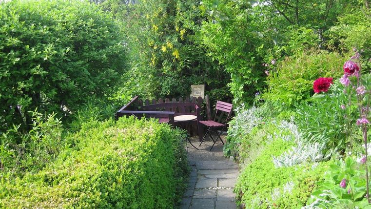 Een mooie tuinpad met dicht groen struikgewas. Op een klein terras staan een tafel en twee stoelen.
