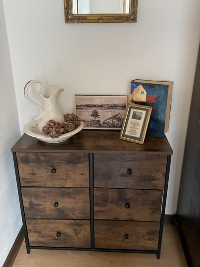Een elegante houten kast met zes lades staat tegen de muur. Daarop staan een kan, een afbeelding, een ingelijst document en enkele decoratieve elementen.