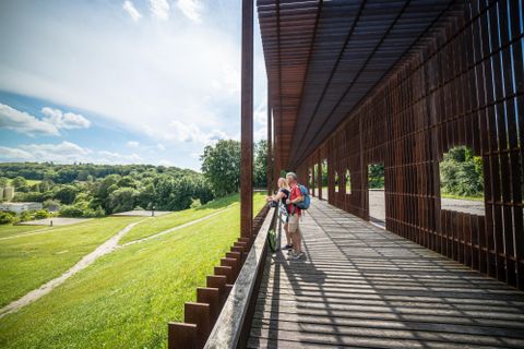 A modern pavilion with a wooden veranda and patterns of shade. Two people enjoy the view of the green landscape and the blue sky.