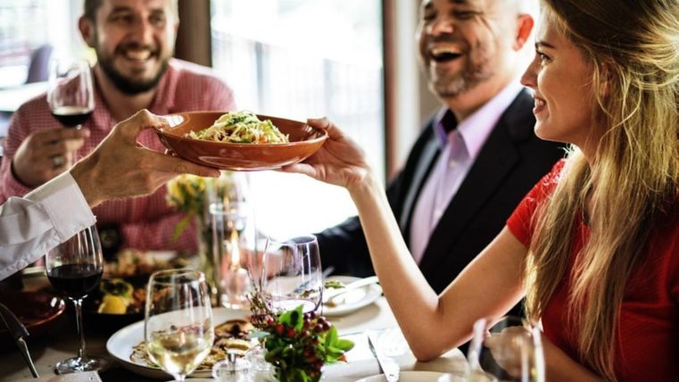 Een groep mensen zit in een restaurant, lacht en geniet van een gezamenlijke maaltijd. Een bord pasta wordt overhandigd.