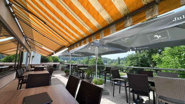 Eine sonnige Terrasse mit gestreiften Markisen und Tischen aus Holz. Der Blick auf das grüne Umland lädt zum Verweilen ein.