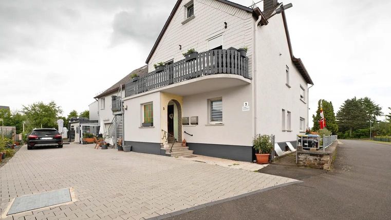 Een modern huis met een overdekt balkon en een geplaveide oprit. De omgeving is groen en goed onderhouden.