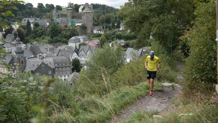 A hiker on a path overlooking a picturesque town with a castle in the background. The surroundings are green and hilly.