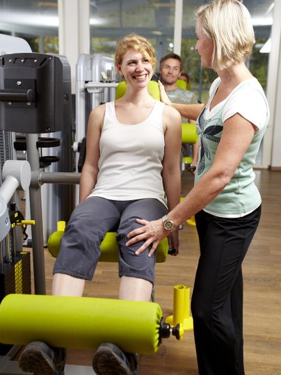 Een vrouw traint op een fitnessapparaat en lacht. Een trainster geeft haar instructies en ondersteuning.