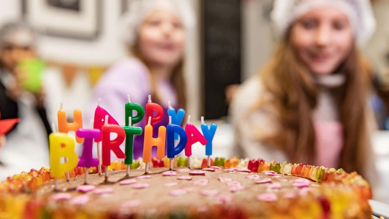Ein Geburtstagskuchen mit bunten Kerzen und Zuckerstreuseln. Im Hintergrund sind zwei Mädchen zu sehen, die fröhlich feiern.
