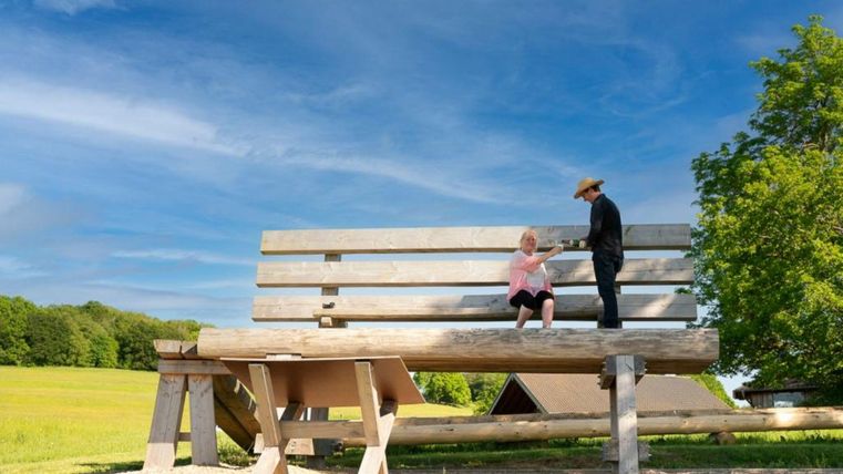Een grote houten bank staat in een groene landschap onder een blauwe lucht. Twee figuren zijn op de bank; de een zit en de ander staat ernaast.