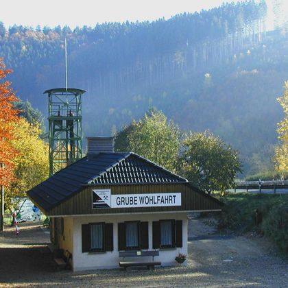 A building complex with the sign "Grube Wohlfahrt". In the background, colorful trees and gentle hills can be seen.