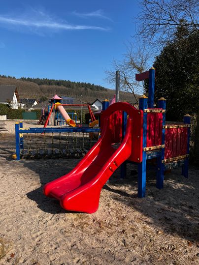 A colorful playground with a red slide and a climbing structure. In the background, other play equipment and a blue sky can be seen.
