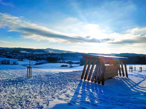 Een winterlandschap met met sneeuw bedekte velden en een houten schuilplaats. De zon schijnt over de heuvels en werpt lange schaduwen.