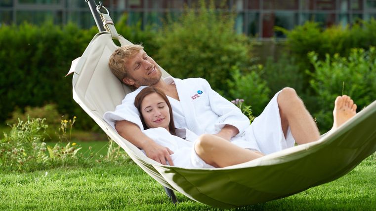 Een stel ontspant in een hangmat op een groen weiland. Beide dragen witte badjassen en genieten van de rust van de natuur.