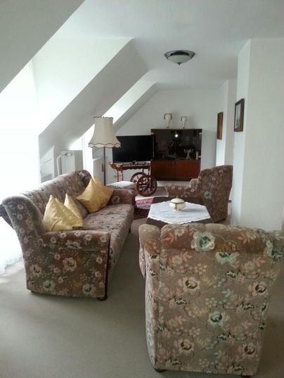 Ein gemütliches Wohnzimmer mit blühenden Sofas und farbigen Kissen. Der Raum ist hell und einladend, mit einem Tisch und einem Fernseher im Hintergrund.