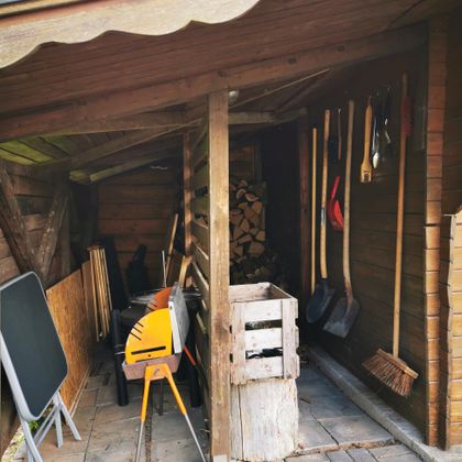 Een kleine tuinhuis met gereedschap aan de muur en houtstapels. Op de grond staan verschillende gereedschappen en een houten kist.