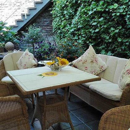 Eine gemütliche Terrasse mit Rattanmöbeln und bunten Kissen. Auf dem Tisch steht eine Vase mit Sonnenblumen und es sind viele Pflanzen im Hintergrund.