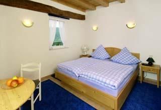 Ein gemütliches Schlafzimmer mit einem großen Doppelbett und blauen Kissen. Der Raum hat einen kleinen Tisch mit Früchten und ein Fenster, das Licht hereinlässt.