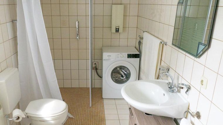 Ein modernes Badezimmer mit einer Dusche, einer Toilette und einem Waschbecken. Eine Waschmaschine ist ebenfalls vorhanden, und die Wände sind mit hellen Fliesen gefliest.