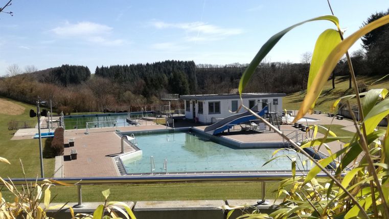 Ein Schwimmbad mit mehreren Becken und einer Rutsche in einer ruhigen Umgebung. Im Hintergrund sind die sanften Hügel und der blaue Himmel zu sehen.