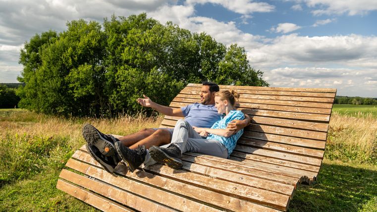 Ein Paar sitzt entspannt auf einer modernen Holzliege in einer grünen Wiese. Im Hintergrund sind Bäume und ein blauer Himmel mit Wolken zu sehen.