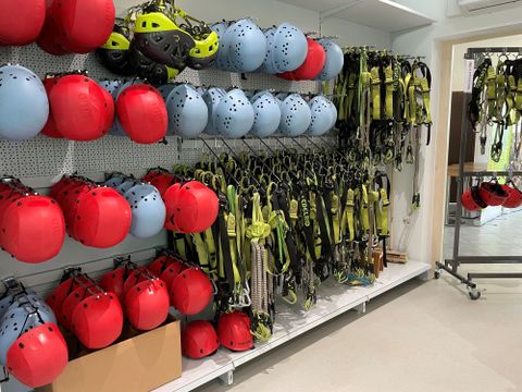 Eine Wand mit mehreren Helmen in Blau und Rot sowie Sicherheitsgeschirren in Gelb. Die Ausrüstung ist ordentlich auf Regalen und Ständern angeordnet.