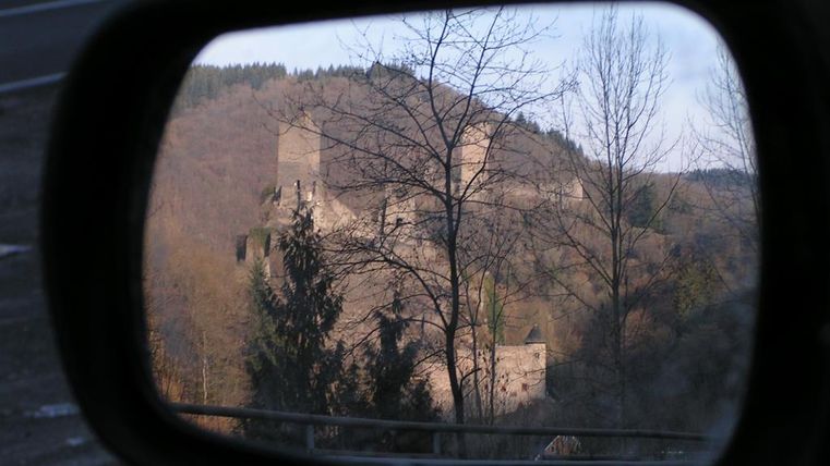 Eine alte Burgruine auf einem Hügel, gesehen im Rückspiegel eines Autos. Die Landschaft ist von Bäumen umgeben und die Atmosphäre ist ruhig.